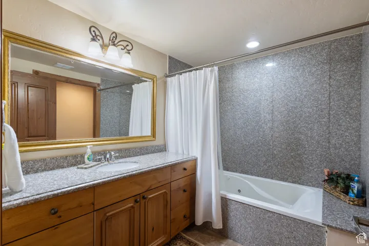 Bathroom featuring a gold-framed vanity mirror, a single basin sink set into a speckled countertop, natural wood-finish cabinetry, a white soaking tub, and speckled wall paneling surrounding the tub