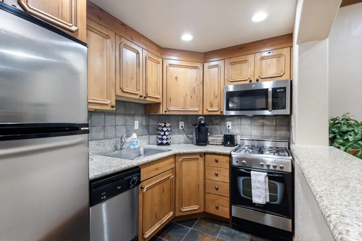 Compact kitchen featuring stainless steel appliances, light wood cabinetry, stone-look countertops, a tiled backsplash, and slate-style floor tiling