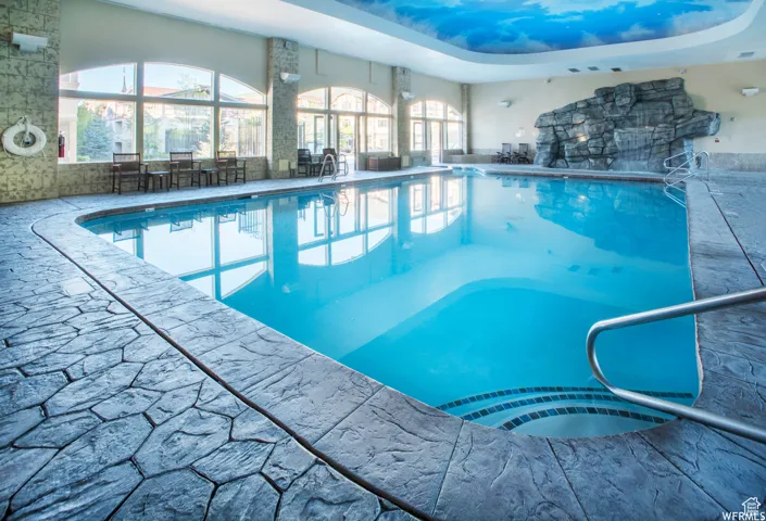 Indoor swimming pool featuring a decorative rock wall, arched windows, and a cloud-mural ceiling