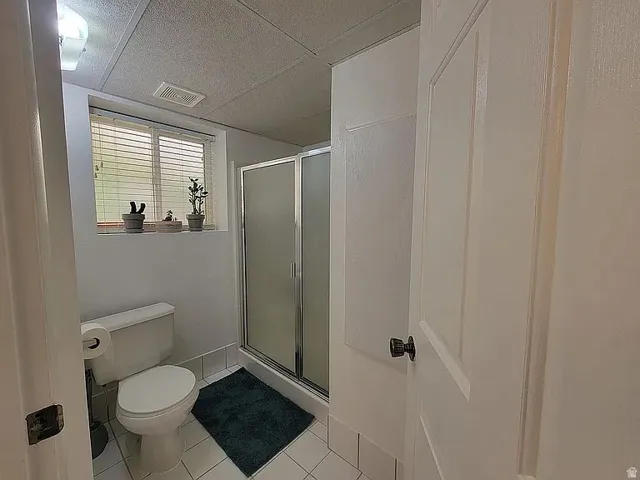 Bathroom featuring a white toilet, enclosed shower with frosted glass, tiled flooring, and a window with blinds
