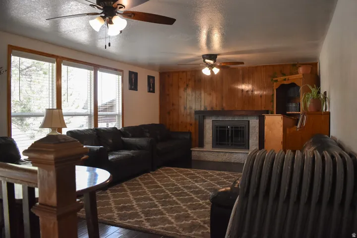 Living area featuring wood-finish flooring, a stone-surround fireplace with a dark wood mantel, wood paneling, and multiple windows with blinds