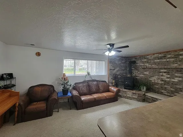 Living area featuring a brick accent wall with a wood-burning stove and hearth, ceiling fan, and carpet flooring