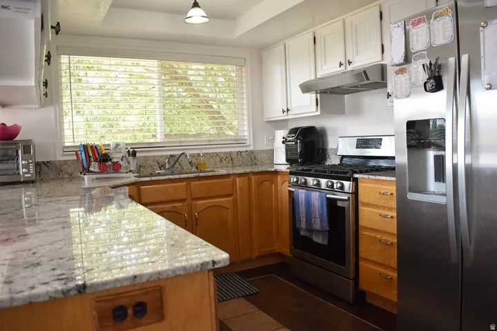 Kitchen featuring granite countertops, a stainless steel range, a stainless steel refrigerator, white upper cabinetry, and natural wood-finish lower cabinetry