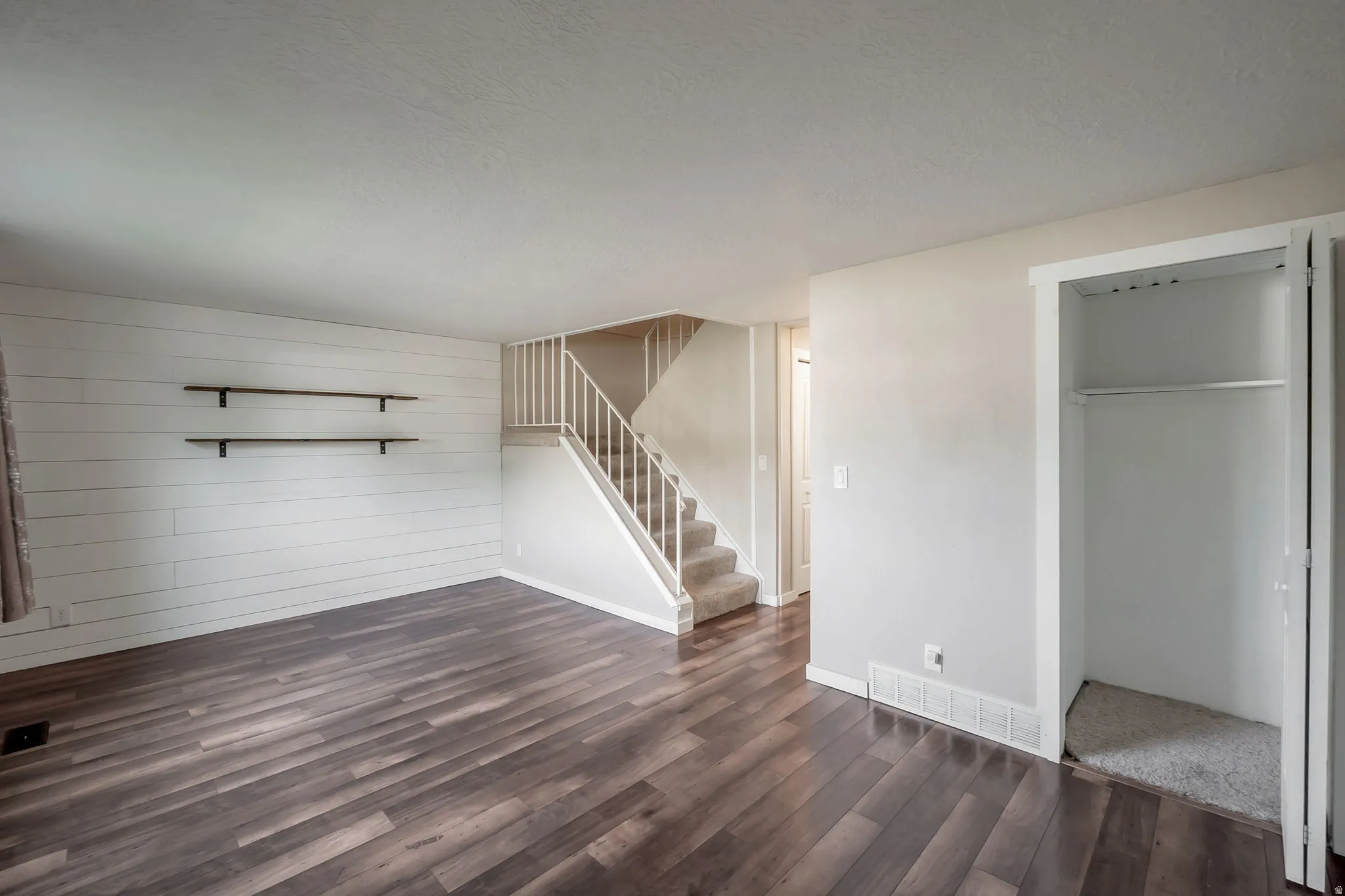 Spacious interior featuring wood-finish flooring, a decorative shiplap accent wall with two floating shelves, a carpeted staircase with white railings, and a coat closet with bi-fold doors