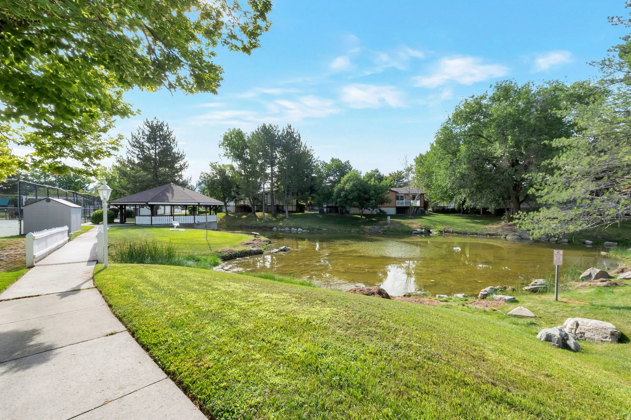 Community pond with a landscaped bank, a nearby gazebo, a concrete pathway, a pedestrian bridge, and established trees