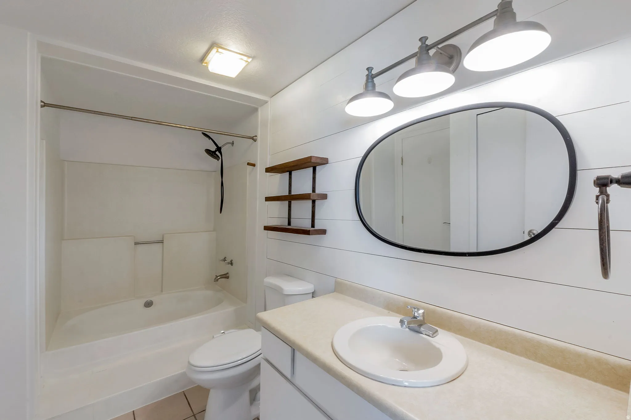 Bathroom featuring shiplap accent wall, white tub/shower combination, oval vanity mirror, wood-finish floating shelves, and a white vanity with a single sink