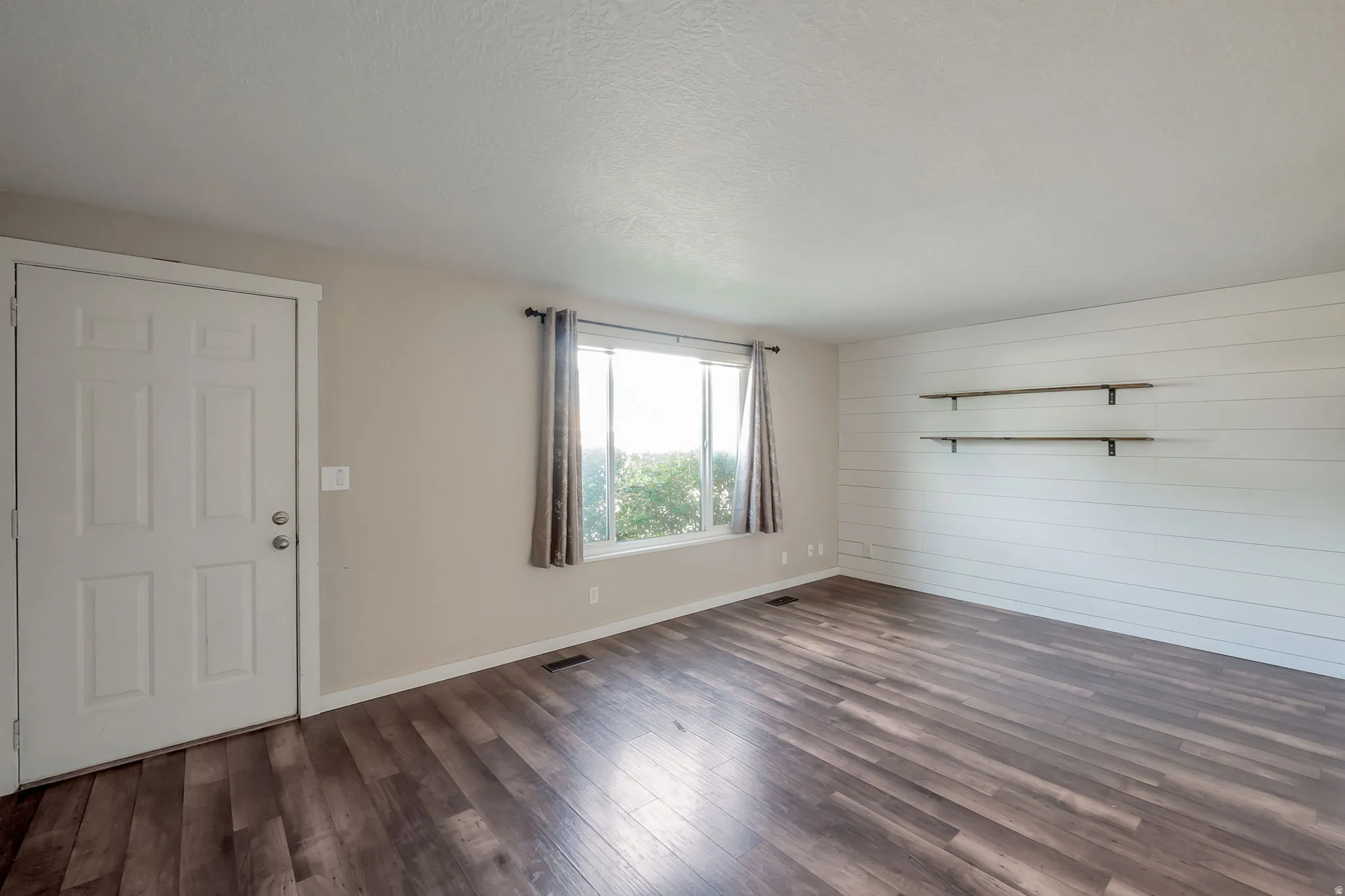 Spacious room featuring wood-finish flooring, a bright window with curtains, and a white six-panel entry door