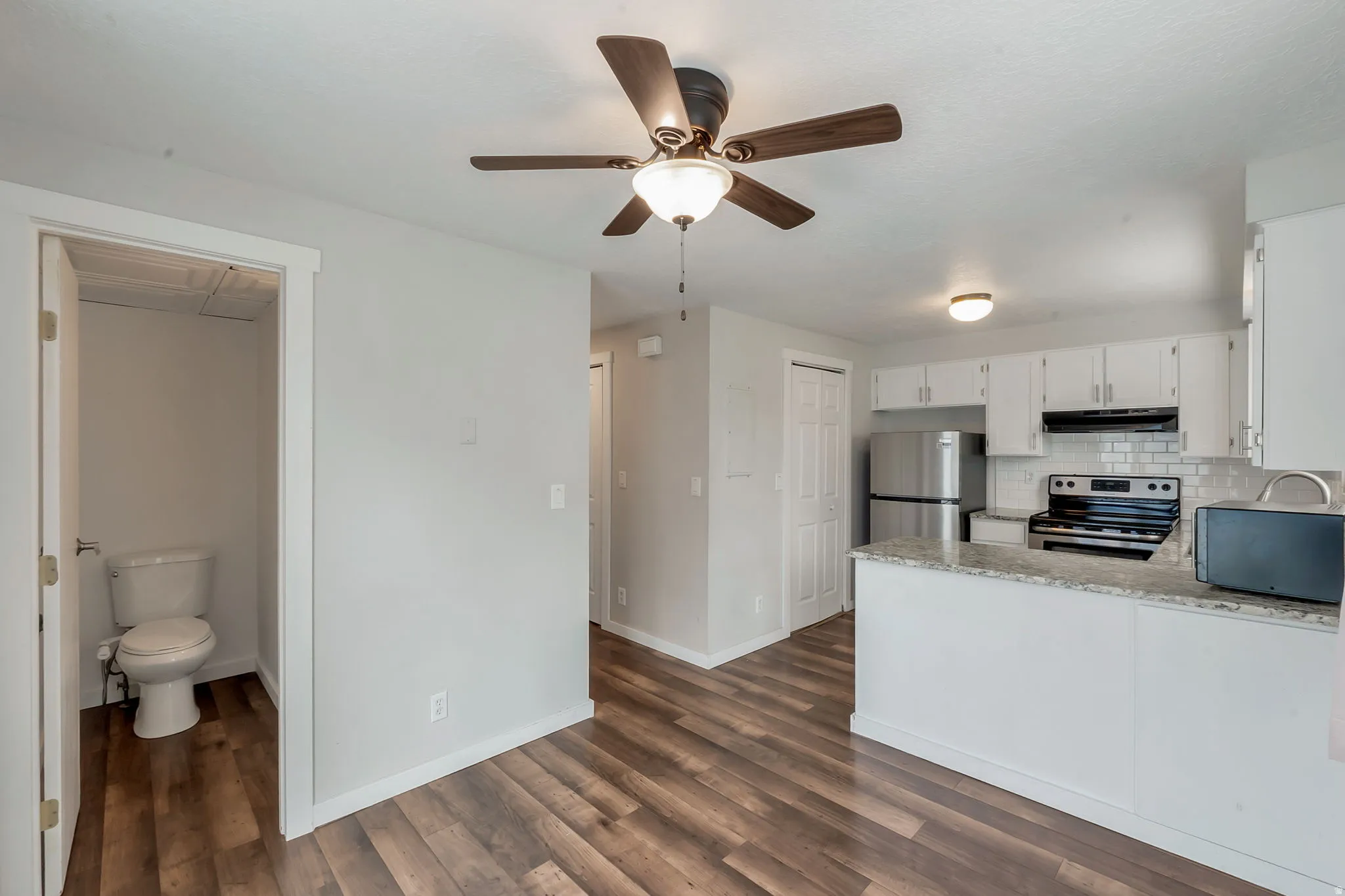 Open-concept living space featuring wood-finish flooring, a ceiling fan with integrated lighting, and a kitchen with white cabinetry, subway tile backsplash, and stainless steel appliances