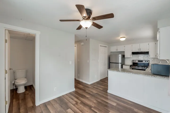 Open-concept living space featuring wood-finish flooring, a ceiling fan with integrated lighting, and a kitchen with white cabinetry, subway tile backsplash, and stainless steel appliances