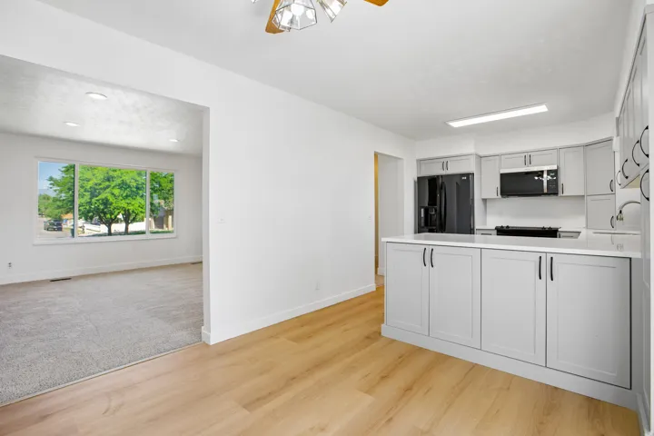 Open-concept living space featuring wood-finish flooring, a kitchen island with light gray shaker cabinetry and white countertops, a suite of black kitchen appliances, and a living area with gray carpeting and a large multi-pane window