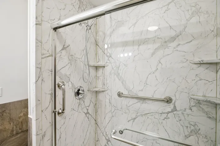 Shower enclosure featuring marble-look wall panels, a chrome shower fixture, a handheld shower head, and built-in corner shelving