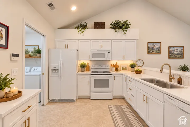 Kitchen featuring vaulted ceilings, white cabinetry with gold-tone hardware, white appliances, dual basin sink with brass-finish faucet, and light-toned tile flooring