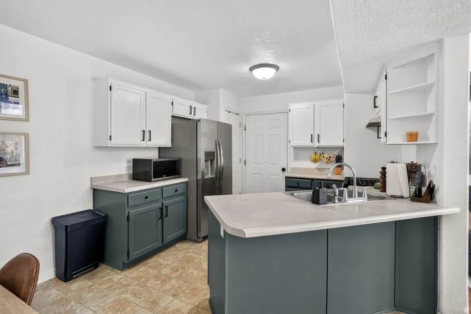 Kitchen featuring two-tone cabinetry with white upper cabinets and deep gray lower cabinets, a stainless steel refrigerator, and a kitchen island with an integrated sink and light-toned countertop