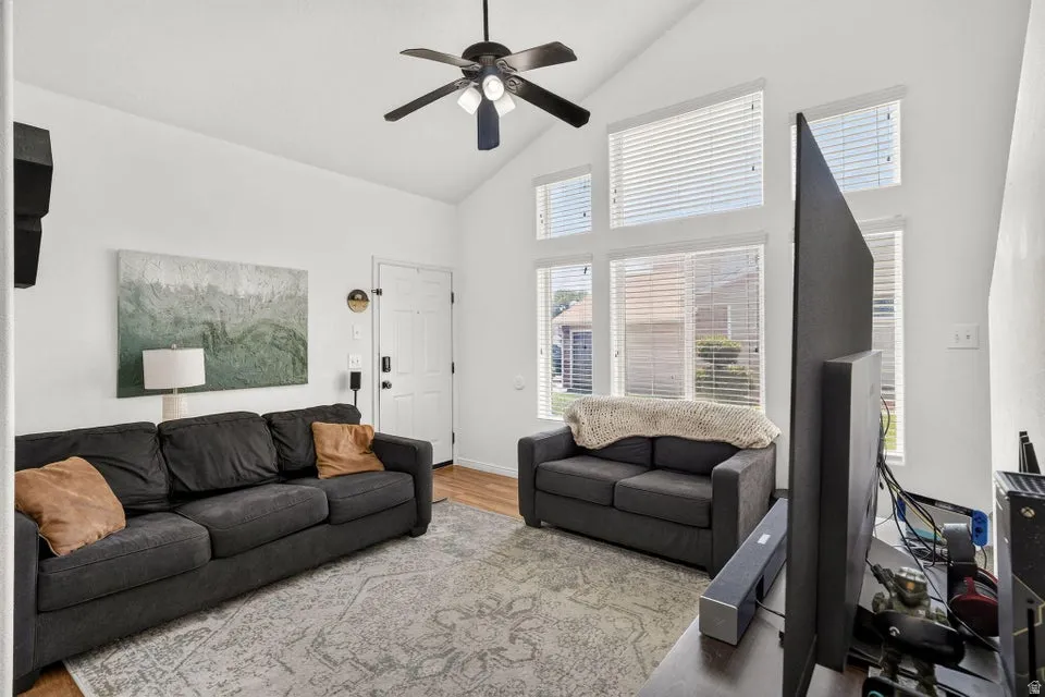 Vaulted ceiling living space featuring a ceiling fan with integrated lighting, wood-finish flooring, a white paneled entry door, and multiple upper windows