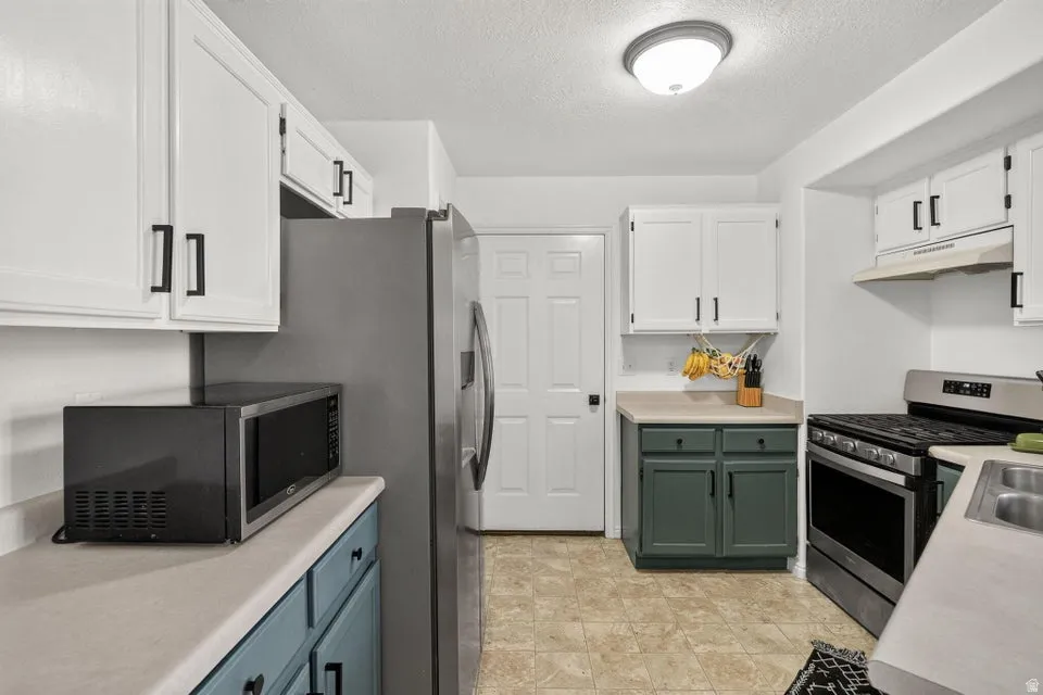 Kitchen featuring white upper cabinetry with black hardware, complementing lower cabinetry in blue and green hues, light-toned countertops, and a stainless steel gas range with a matching range hood