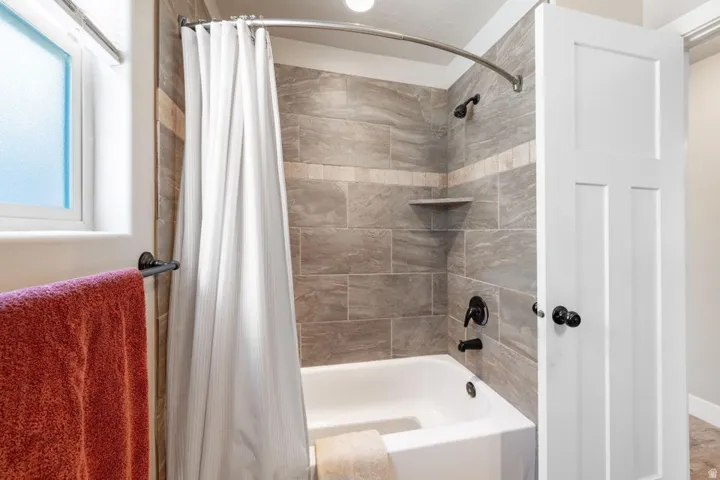 Bathtub and shower combination featuring floor-to-ceiling gray tile surround with a decorative horizontal band, matte black fixtures, and a corner shelf