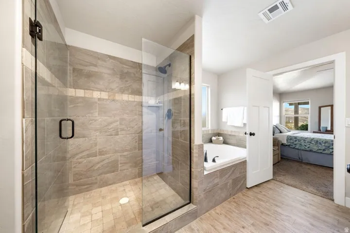 Spacious bathroom featuring a glass-enclosed shower with stone-look tile, a built-in soaking tub, and light wood-finish flooring