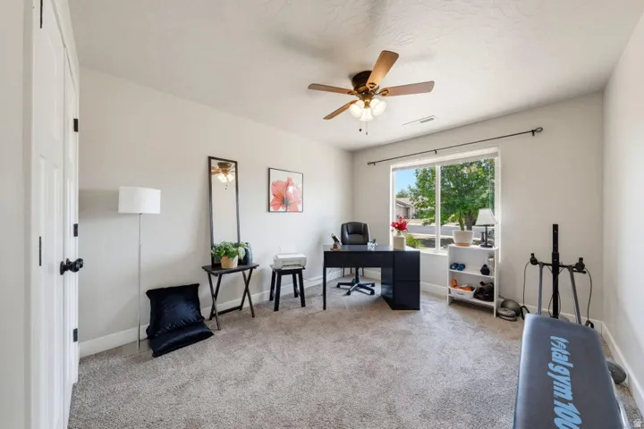 Spacious room featuring light gray carpeting, neutral wall paint, a large window with exterior landscaping, a ceiling fan with integrated lighting, and white trim throughout