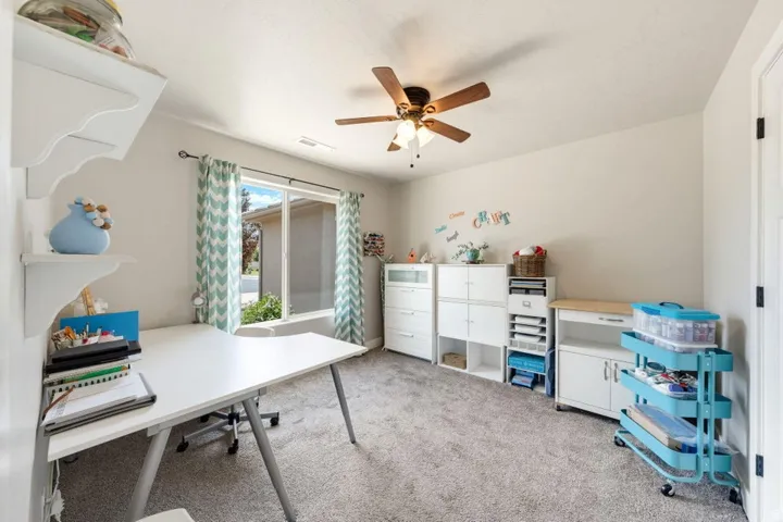 Carpeted room featuring a large window with drapes, ceiling fan with light fixture, and white wall shelving