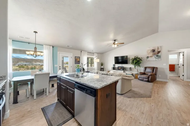 Open concept living area featuring a kitchen island with a double basin sink and granite-style countertop, wood-finish flooring, vaulted ceilings, and a dining nook with a scenic backdrop