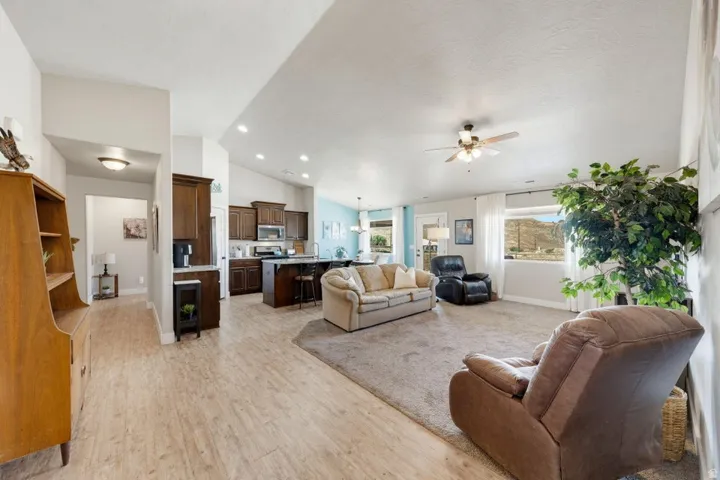 Open-concept living space featuring wood-finish flooring, a kitchen with dark wood cabinetry and an island, a carpeted living area, a ceiling fan, and large windows with outdoor scenery
