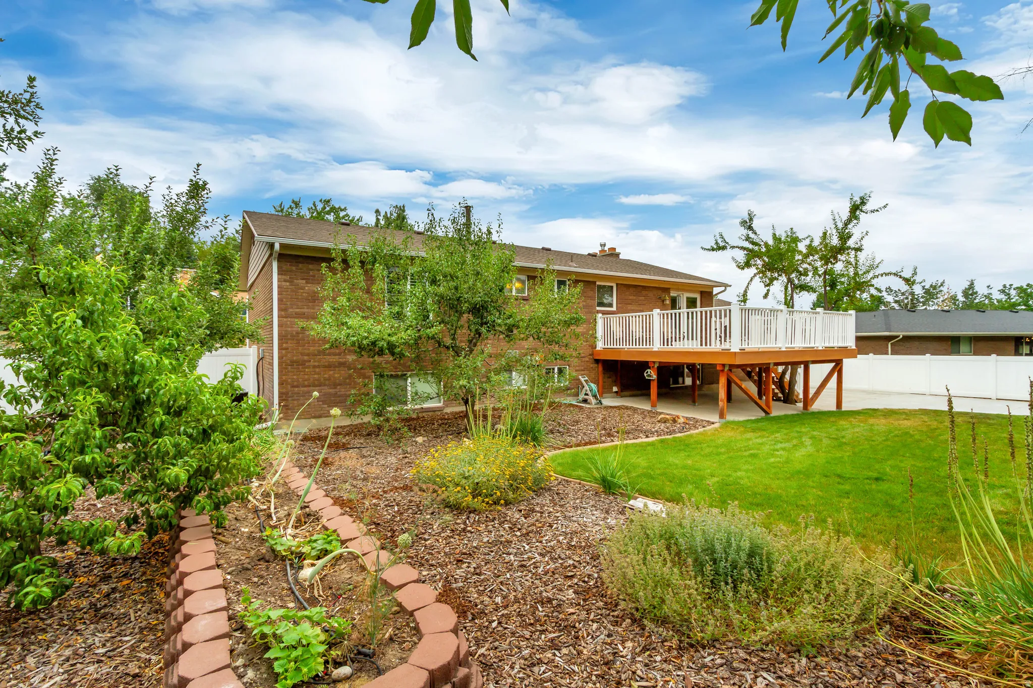 Brick exterior featuring a multi-level deck with white railings, a manicured lawn, and a garden bed bordered by brick pavers