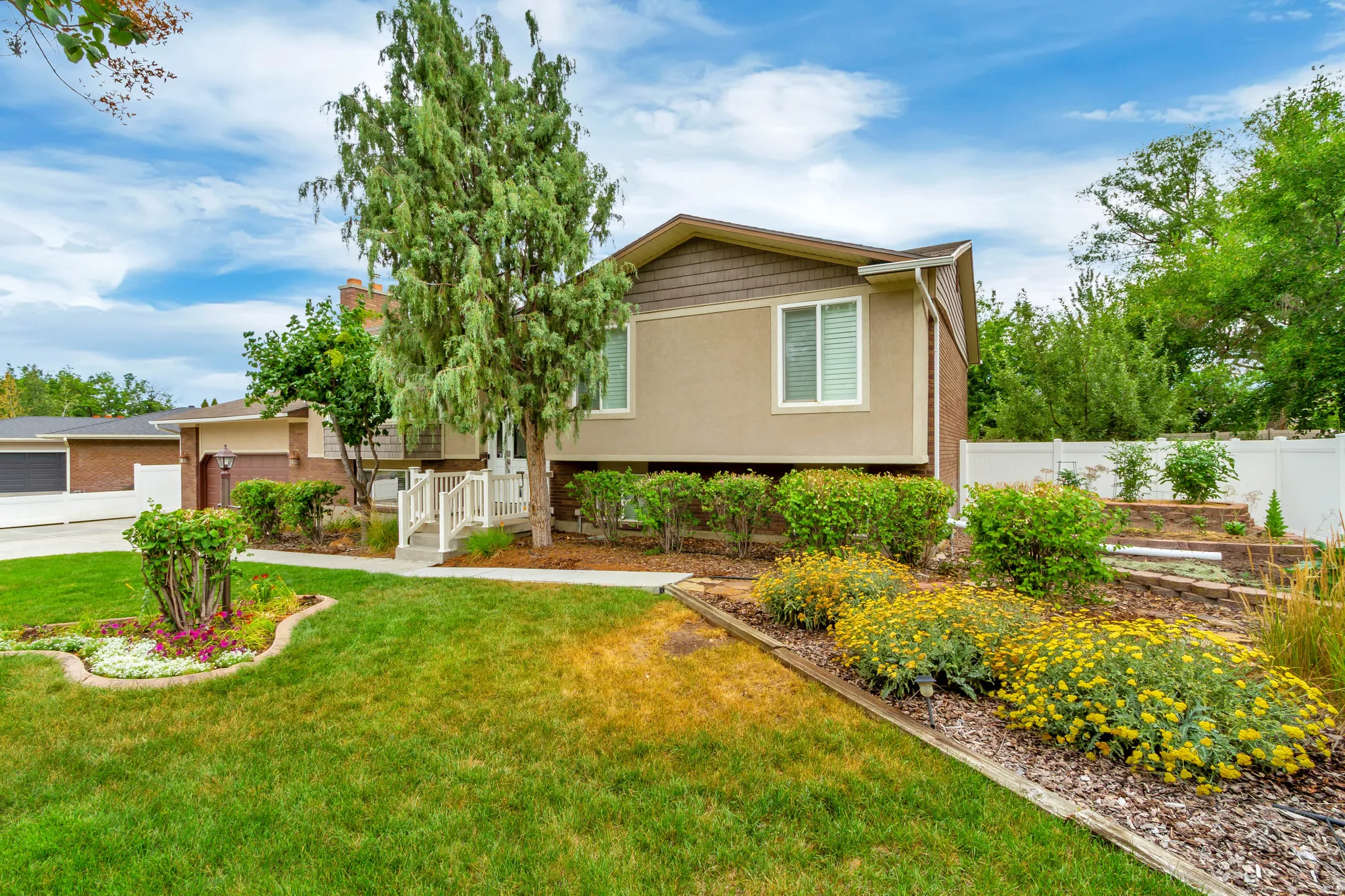 Split-level residence featuring a stucco and brick exterior, white railing entry stairs, a concrete walkway, mature landscaping with flowering shrubs, and a white privacy fence