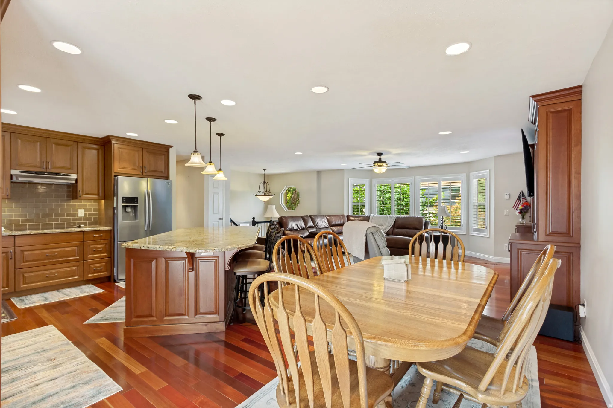Open-concept living area featuring rich wood-finish flooring, a kitchen island with pendant lighting, recessed ceiling lights, and bay windows with plantation shutters