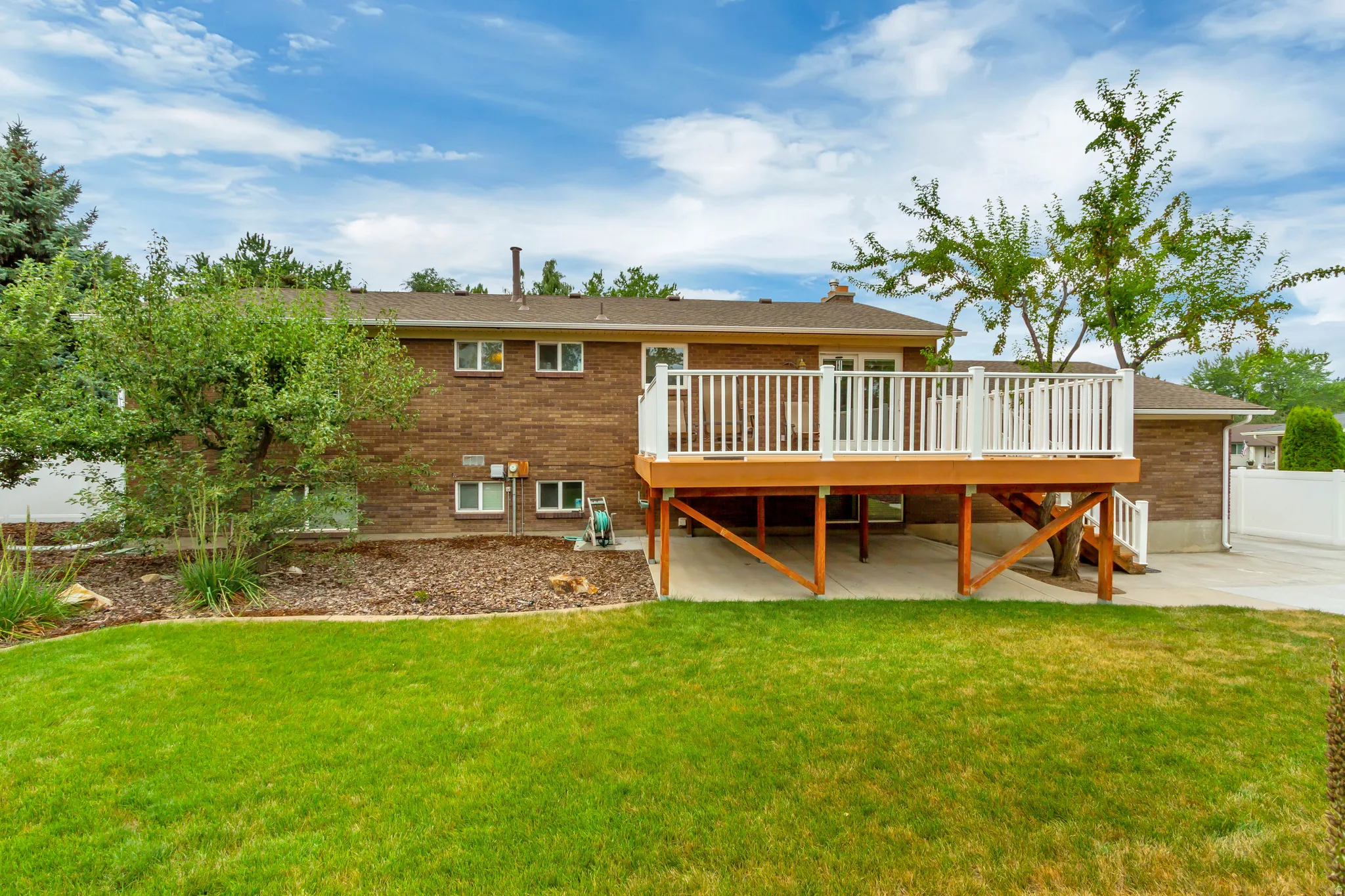 Brick exterior featuring an elevated wood deck with white railings, a concrete patio beneath, and a manicured lawn