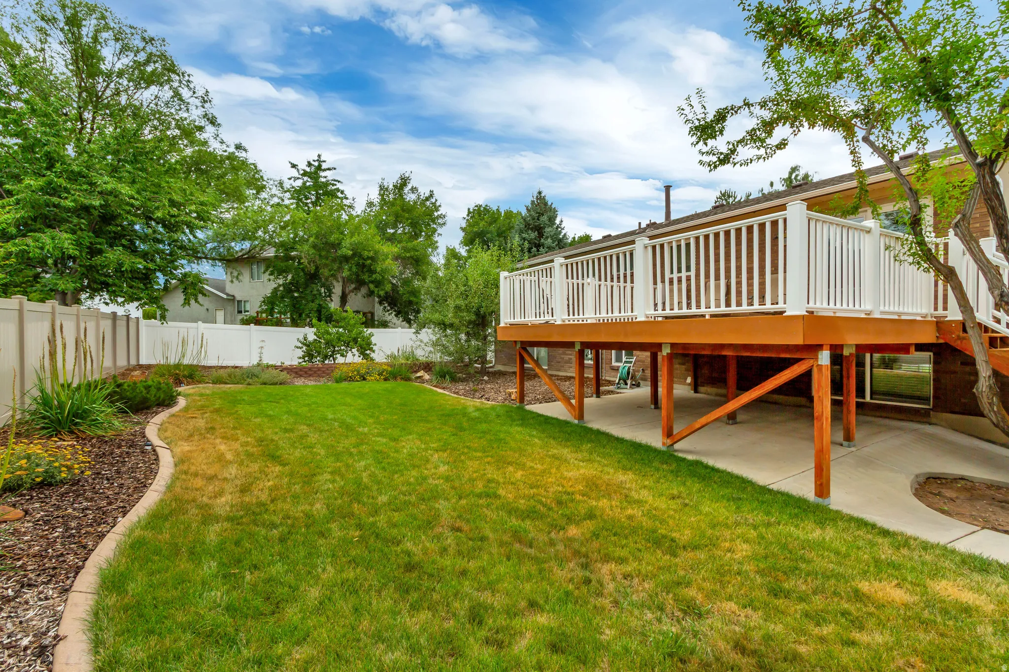 Expansive backyard featuring a large raised deck with white railings, a concrete patio area, a manicured lawn, and landscaped garden beds