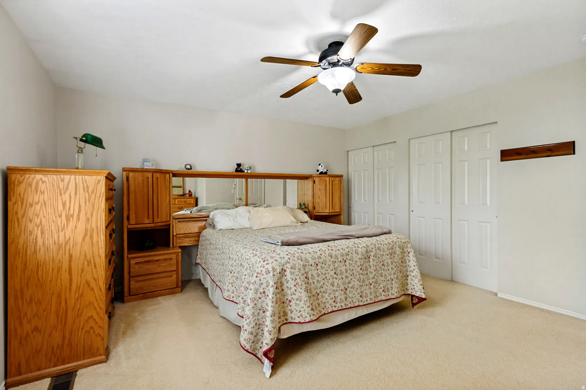 Carpeted room featuring a ceiling fan with integrated lighting, two sliding closet doors, and a wall-mounted wooden rack