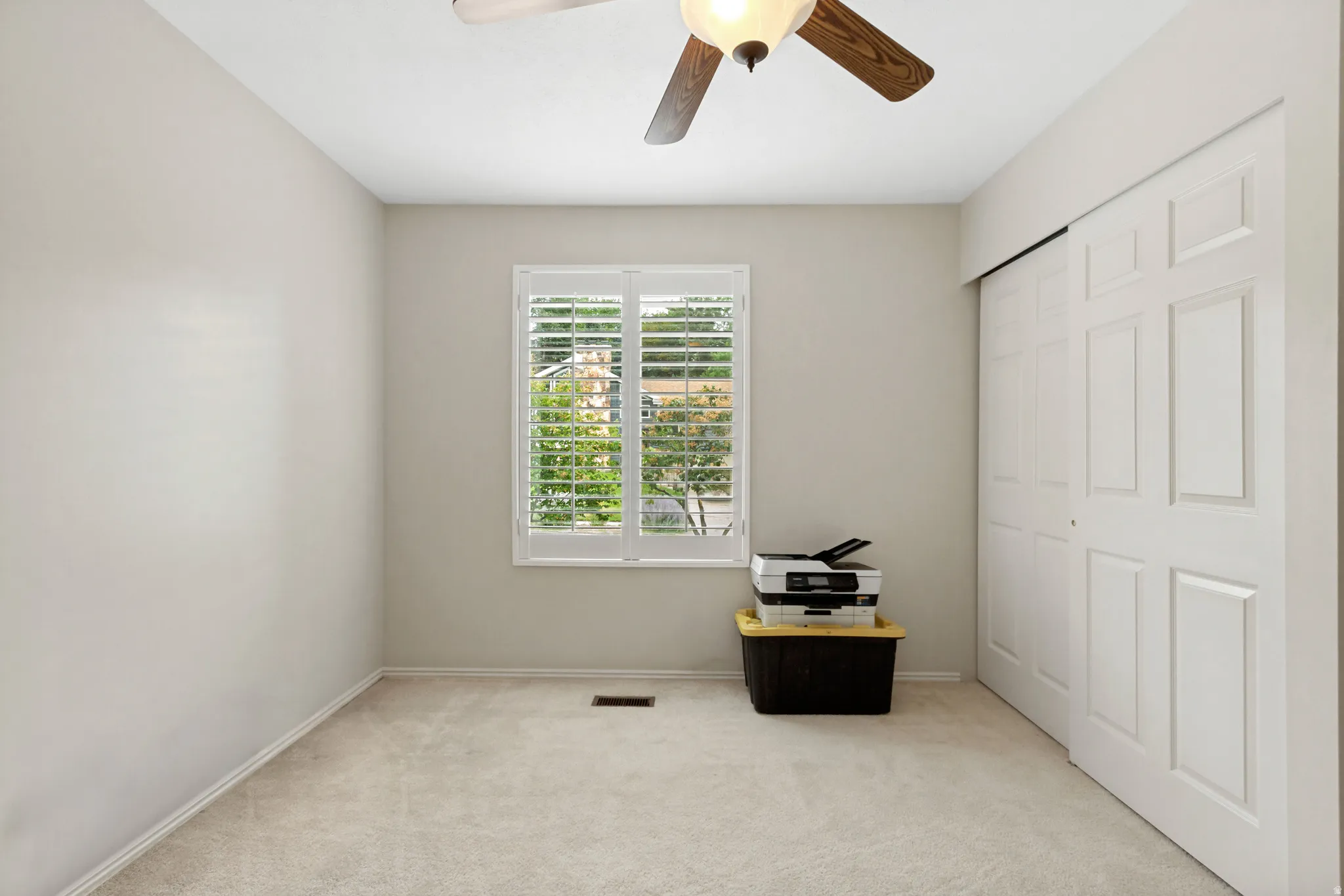 Neutral-toned room featuring a window with plantation shutters, ceiling fan with wood-finish blades, and wall-to-wall carpeting