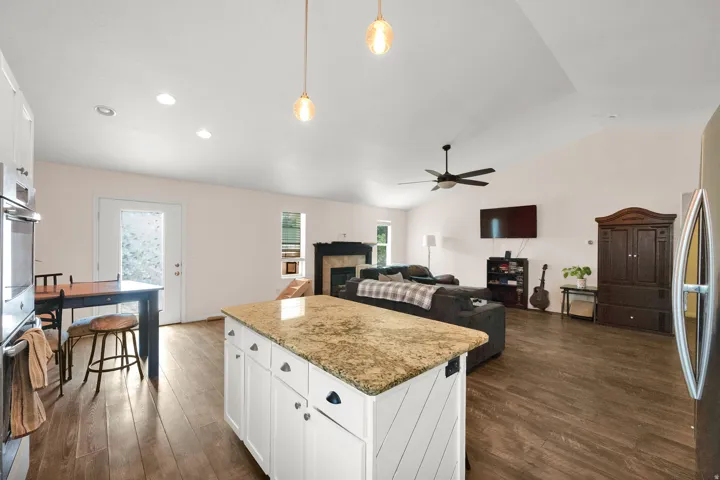 Kitchen island with granite countertop and white cabinetry, integrated into an open living space featuring vaulted ceilings, wood-finish flooring, and a fireplace with a black surround