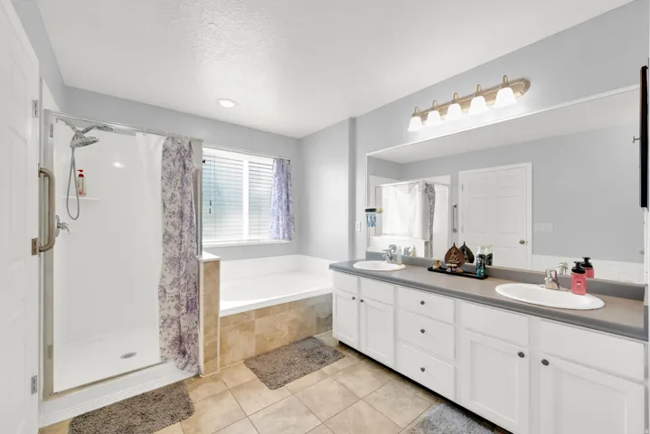 Bathroom featuring a dual vanity with white cabinetry, a large mirror, a separate shower with a glass enclosure, a built-in tub, and tile flooring