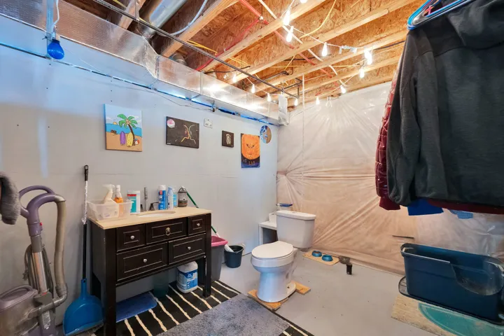 Plumbed for a basement bath, toilet working, to be relocated.  Basement area featuring a dark wood vanity with a light-colored countertop, exposed ceiling joists, string lighting, and light gray painted walls