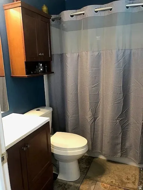 Full bathroom featuring a white toilet, brown wood-finish vanity with a white countertop, and a wall-mounted brown wood-finish cabinet with open shelving