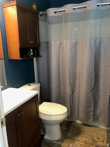 Full bathroom featuring a white toilet, brown wood-finish vanity with a white countertop, and a wall-mounted brown wood-finish cabinet with open shelving