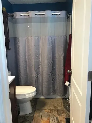 Bathroom with shower, featuring a white toilet, dark wood-finish cabinet with white countertop, and varied-tone tile flooring