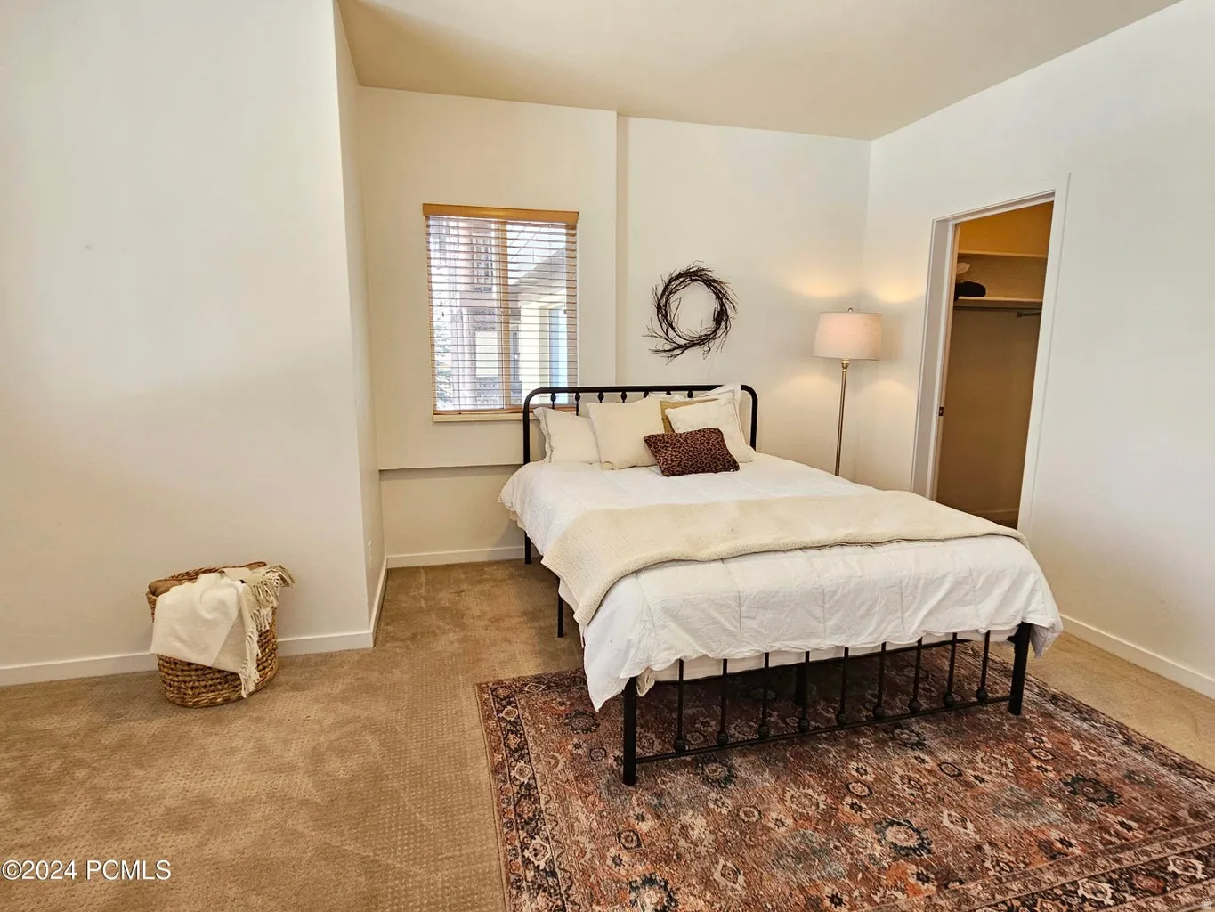 Carpeted room with a window featuring wood-slat blinds, a metal bed frame, and an open doorway revealing a closet with shelving