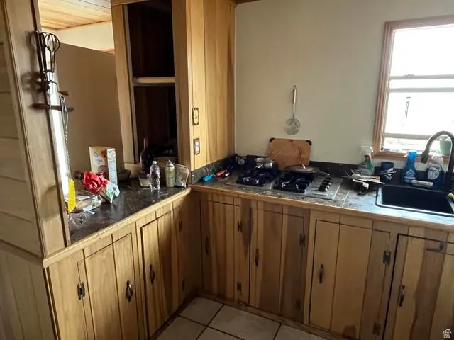 Kitchen featuring natural wood cabinetry, a built-in cooktop, a dark-toned sink with a gooseneck faucet, a window providing natural light, and tile flooring