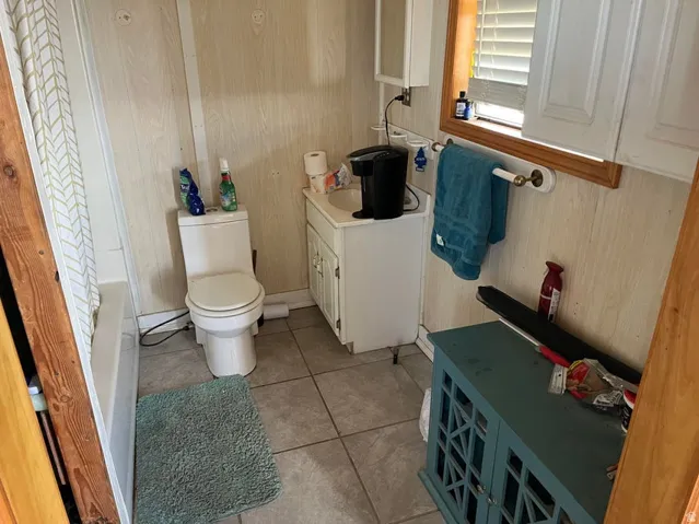 White paneled walls, floor tiling, white vanity with integrated sink, white toilet, and window with blinds