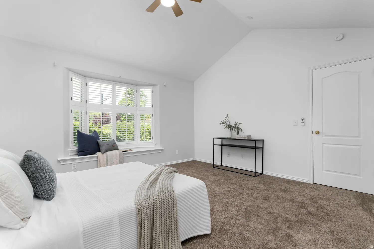 Primary Bedroom~Spacious room featuring vaulted ceilings, a prominent bay window with white plantation shutters, a ceiling fan, and plush carpeting
