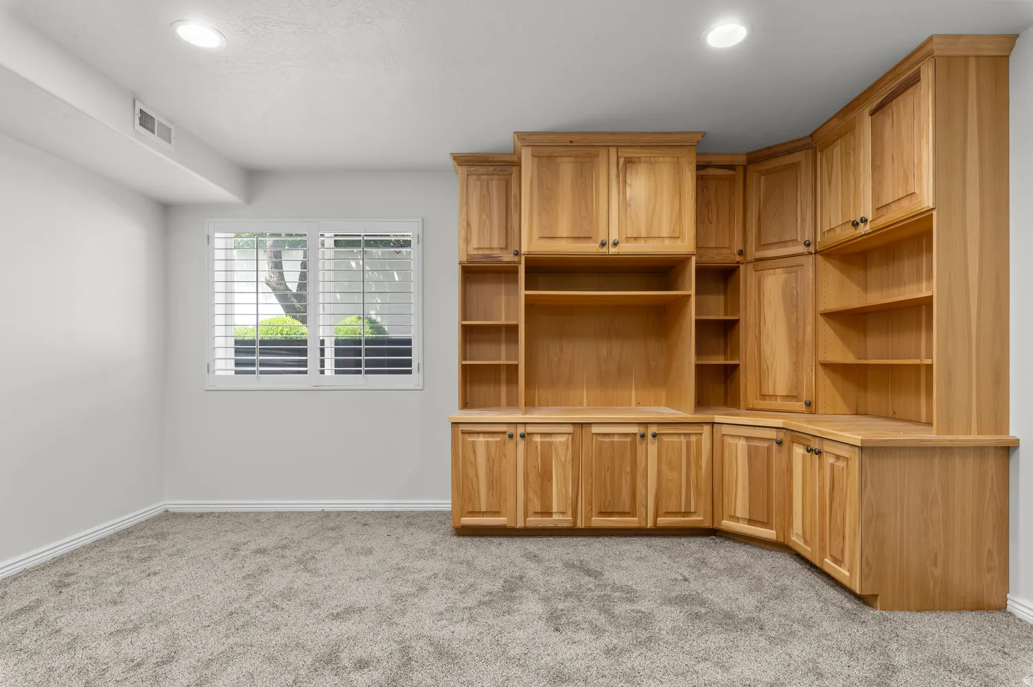 Built-in wood cabinetry with integrated shelving, recessed lighting, double pane window with white plantation shutters, light gray wall paint, and gray carpet flooring