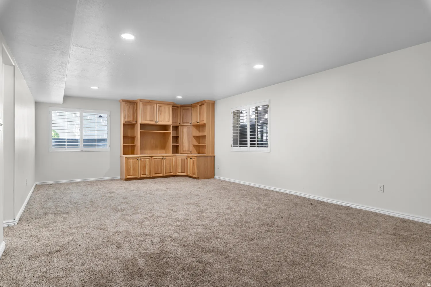 Spacious room featuring a corner built-in entertainment center with natural wood cabinetry and shelving, plush carpeting, recessed lighting, and two windows with white louvered blinds