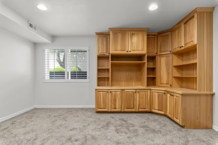 Built-in wood cabinetry with integrated shelving, recessed lighting, double pane window with white plantation shutters, light gray wall paint, and gray carpet flooring