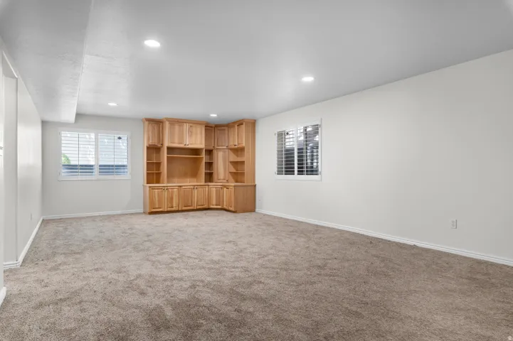 Spacious room featuring a corner built-in entertainment center with natural wood cabinetry and shelving, plush carpeting, recessed lighting, and two windows with white louvered blinds