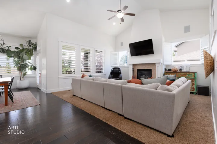 Vaulted ceilings highlight this living space, featuring a fireplace with a mantel, multiple windows with plantation shutters, dark wood-finish flooring, and a ceiling fan