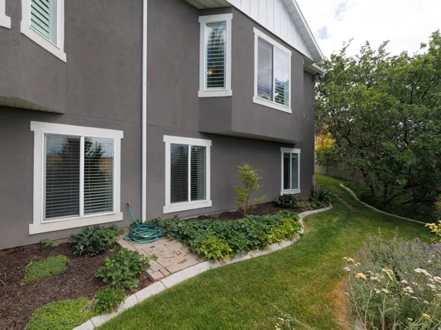 Gray stucco exterior featuring white-framed windows, a landscaped planting bed, mature trees, and a manicured lawn