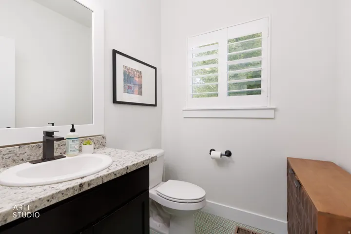 Bathroom featuring a white framed mirror, a vanity with a light-colored speckled countertop, a white oval sink with a black faucet, a window with white plantation shutters, and light green mosaic floor tiling