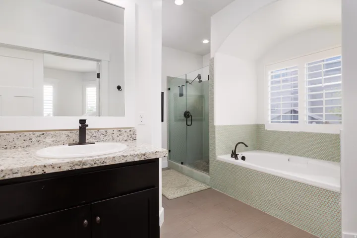 Bathroom featuring a built-in soaking tub with light green penny tile surround, a glass-enclosed shower, a vanity with a speckled countertop, an oval undermount sink, and a framed mirror
